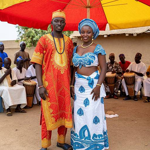 Photograph of two African couples standing under a red and yellow umbrella, wearing traditional bright orange and blue patterned outfits, with drummers in the background