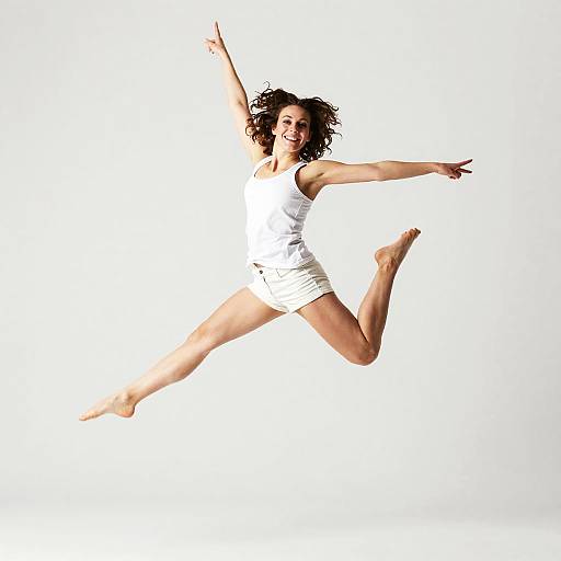 Photograph of a smiling woman with curly dark hair, wearing a white tank top and shorts, mid-air jump against a white background.