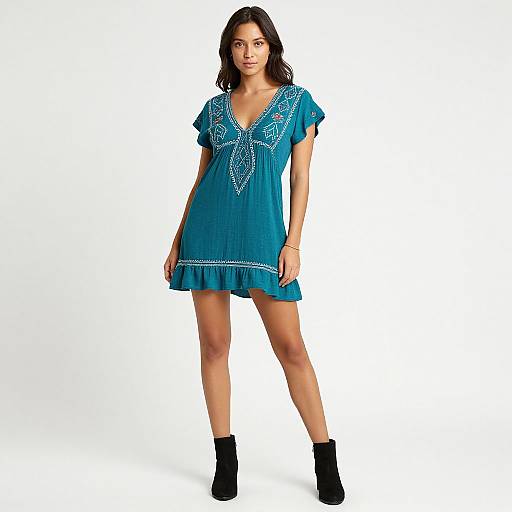 Photograph of a young woman with long dark hair, wearing a turquoise short-sleeved dress with white embroidery, black ankle boots, standing against a