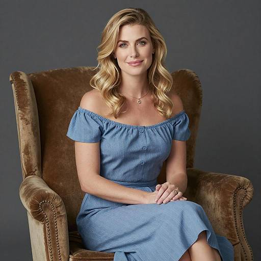 Photograph of a blonde woman with wavy hair, wearing an off-shoulder blue dress, seated in a brown velvet armchair against a dark