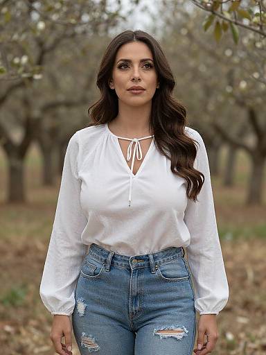 Photograph of a dark-haired woman with wavy hair, wearing a white long-sleeve blouse and ripped blue jeans, standing in an orchard