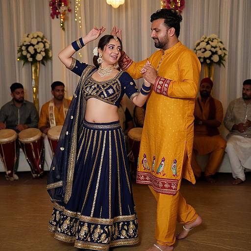 Indian couple dancing in traditional attire; woman in black and gold lehenga, man in yellow kurta. Background: drummers, floral decorations. Photograph