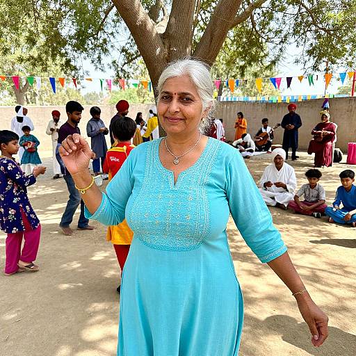 Photograph of an older Indian woman with white hair, wearing a light blue embroidered dress, smiling and waving under a large tree, surrounded by children and