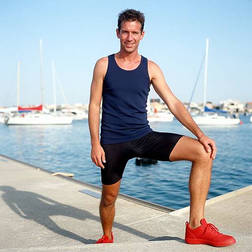 Photograph of a fit, smiling man with short dark hair, wearing a black tank top, black shorts, and red sneakers, posing with one knee