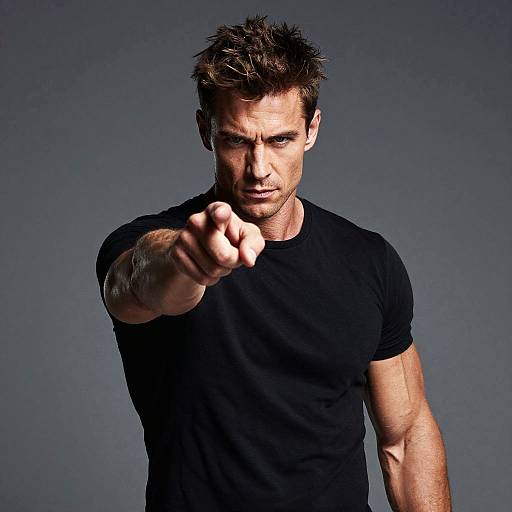 Photograph of a muscular, serious-looking man with spiked brown hair, wearing a black t-shirt, pointing forward against a dark gray background.