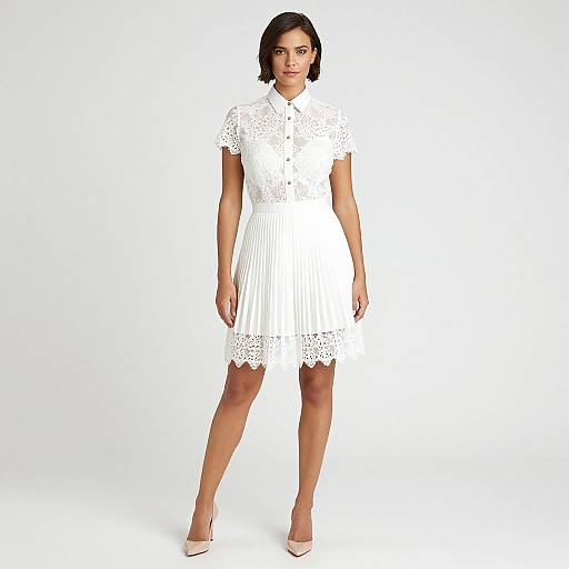 Photograph of a woman with short black hair wearing a white, short-sleeve, lace and pleated dress, standing against a plain white background