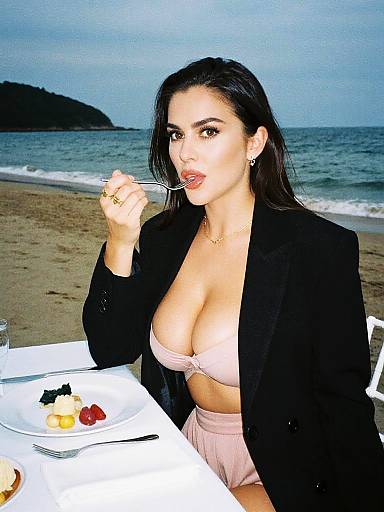 Photograph of a dark-haired woman with large breasts, wearing a black blazer and pink bikini top, eating at a beach table with a plate of