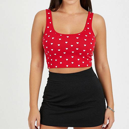 Photograph of a woman with light skin, wearing a red heart-patterned crop top and black high-waisted skirt, against a plain white background