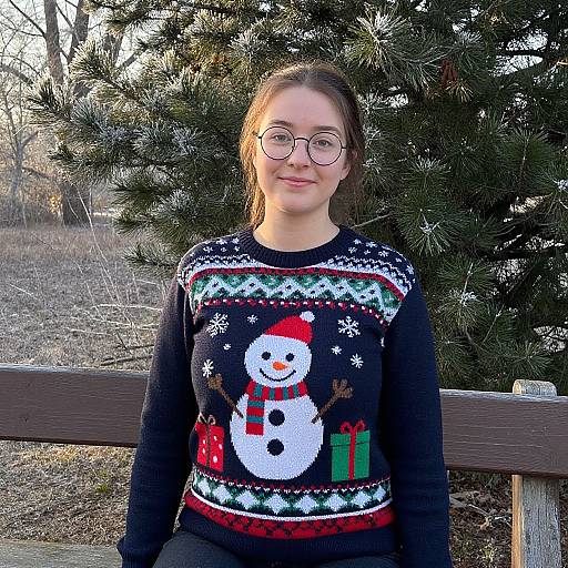 Photograph of a smiling woman with glasses, brown hair, wearing a black Christmas sweater with a snowman and gifts, standing in front of a pine