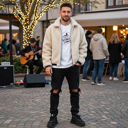 Photograph of a bearded man in a fluffy white jacket, white graphic tee, ripped black jeans, and black boots, standing on a cobble
