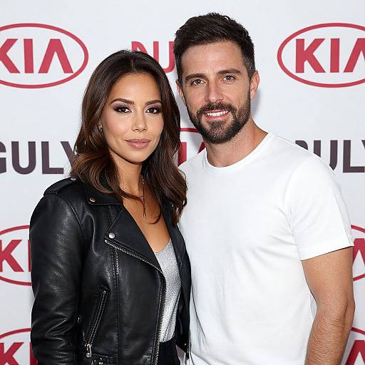 Photograph of a smiling brunette woman in a black leather jacket and white top, standing beside a bearded man in a white t-shirt, against a