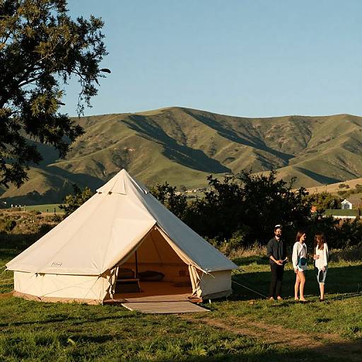 Photograph of a white canvas tent in a grassy field with three people standing nearby, mountainous terrain in the background.