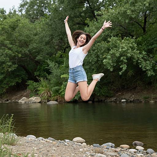 Photograph of a joyful young woman with short dark hair, wearing a white tank top and blue denim shorts, jumping mid-air above a rocky riverbank