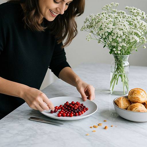 Photograph of a smiling woman with brown hair, wearing a black top, serving vibrant red cranberries from a white plate, with a vase of white