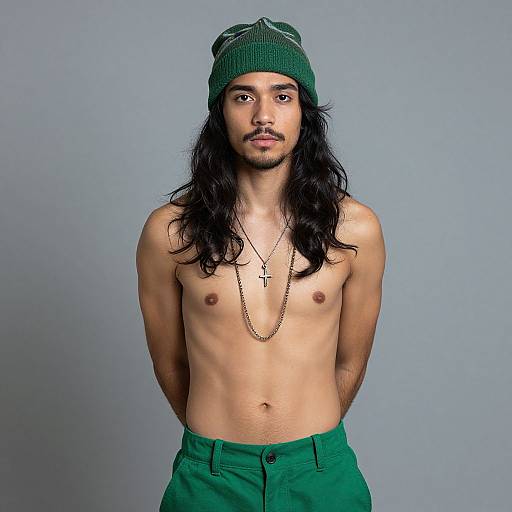 Photograph of a shirtless young man with long black hair, green beanie, green pants, cross necklace, and hands behind back, against a