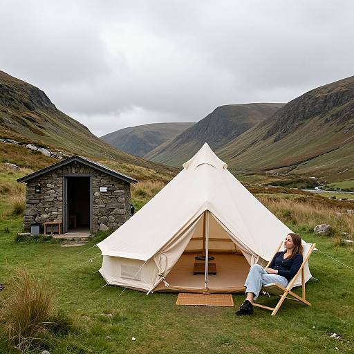 Photograph of a woman in a white camping tent, sitting on a wooden chair, with a stone hut in a mountainous, grassy landscape.