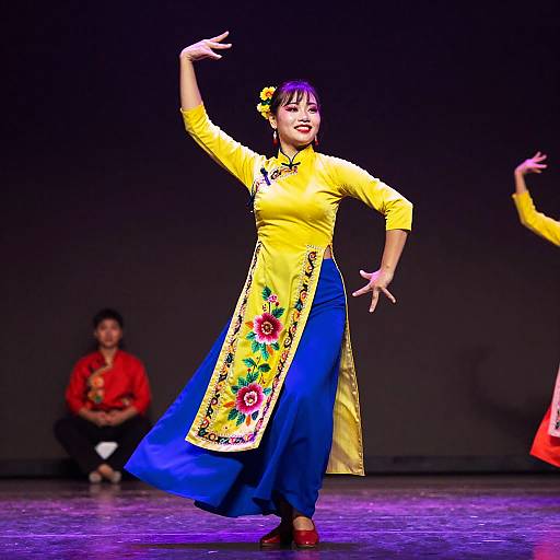 Photograph of an Asian woman in a vibrant yellow and blue traditional dance outfit with floral embroidery, performing on stage with a dark background. Another dancer in