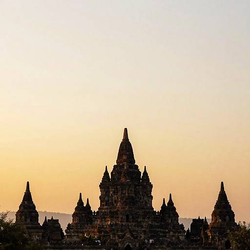 Photograph of a silhouetted ancient Hindu temple complex with multiple pointed spires against a gradient sunset sky, transitioning from yellow to blue.