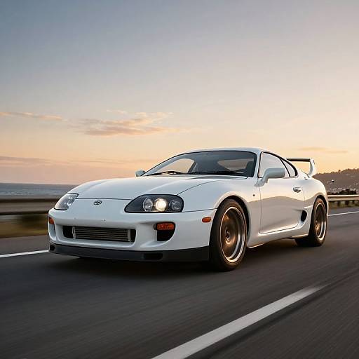 Photograph of a white, modified Toyota Supra racing on a highway at sunset, with its headlights on and a clear sky in the background.