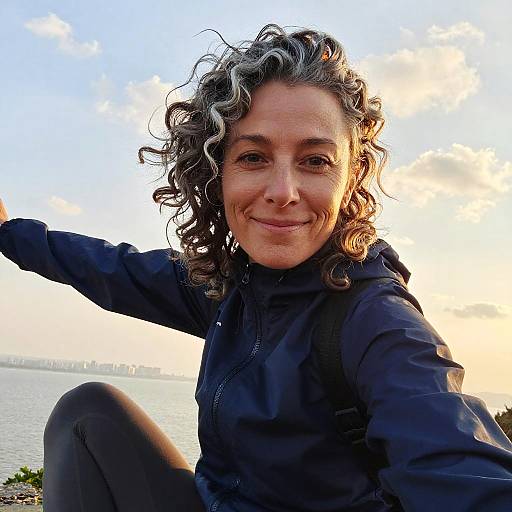 Photograph of a smiling woman with curly gray and brown hair, wearing a black jacket, sitting by a lakeside at sunset.