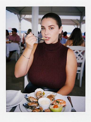 Photograph of a dark-haired woman with fair skin, wearing a sleeveless brown turtleneck, eating a pastry at an outdoor cafe.