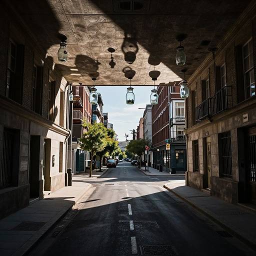 Photograph of a sunlit urban street under a shadowy overhead walkway with hanging lights, flanked by brick buildings and trees.
