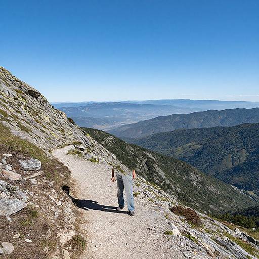 Photograph of a hiker on a rocky mountain trail, with a clear blue sky and expansive mountain range in the background.