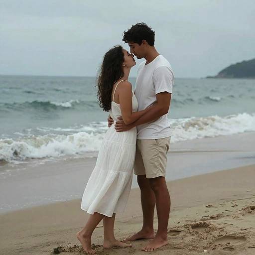 Photograph of a couple kissing on a beach; she wears a white sundress, he wears a white t-shirt and beige shorts. Ocean waves in