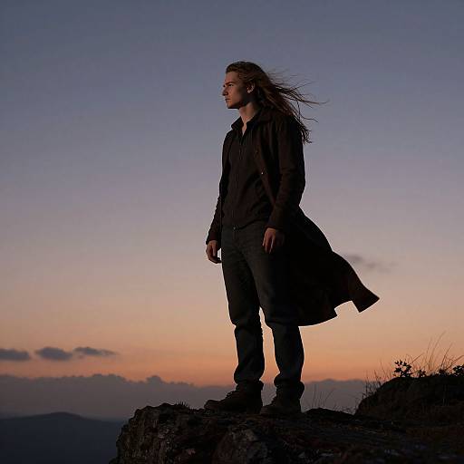Silhouetted man with long hair in dark coat standing on rocky cliff, sunset sky with pink and purple hues in background.