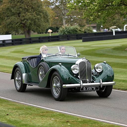 Photograph of a green classic convertible sports car with a white top, driven by a man on a grassy road. Background features trees and a blurred