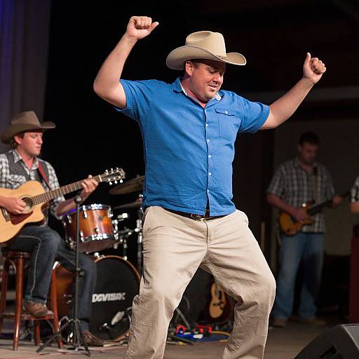 Photograph of a male singer in a blue shirt, beige pants, and cowboy hat energetically performing on stage with three musicians in background.