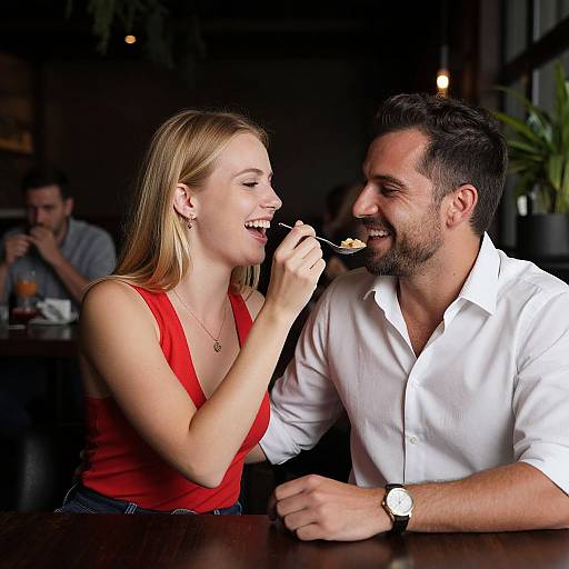 Photograph of a smiling blonde woman in a red sleeveless top and a bearded man in a white shirt feeding her dessert in a dimly lit
