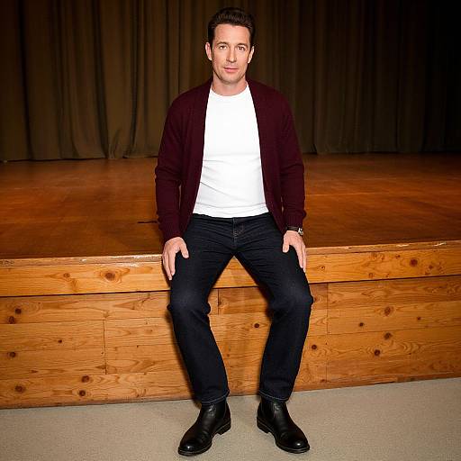 Photograph of a man with short dark hair, wearing a black cardigan, white t-shirt, black jeans, and black boots, sitting on a