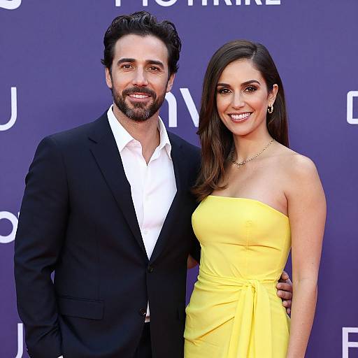 Photograph of a smiling couple at an event: bearded man in black suit and white shirt, woman in strapless yellow dress, blue background.