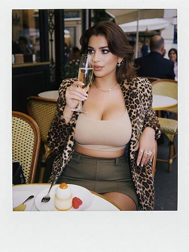 Photograph of a curvy woman with dark hair, wearing a leopard-print jacket, beige top, and green skirt, sipping champagne at a café