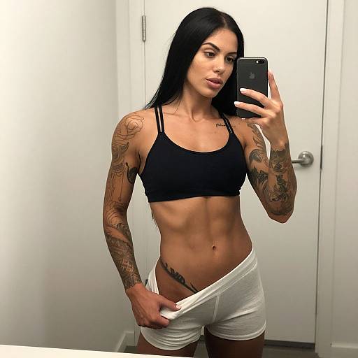 Tattooed woman with long black hair takes a mirror selfie in a white bathroom, wearing a black sports bra and white shorts, showing a toned