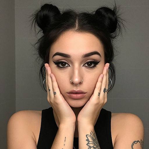 Photograph of a young woman with black hair in double buns, dark makeup, nose ring, hoop earrings, and tattoos, hands framing her face