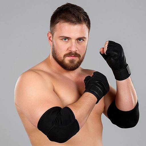 Photograph of a muscular, bearded white man with short brown hair, wearing black fingerless gloves, posing with fists up against a plain gray background