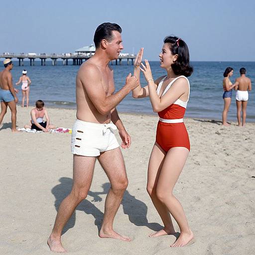 Photograph of a beach scene: a shirtless man in white shorts and a woman in red one-piece swimsuit high-five each other, with other