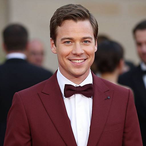 Photograph of a smiling young man with short brown hair, wearing a maroon tuxedo with a black bow tie, in front of a blurred