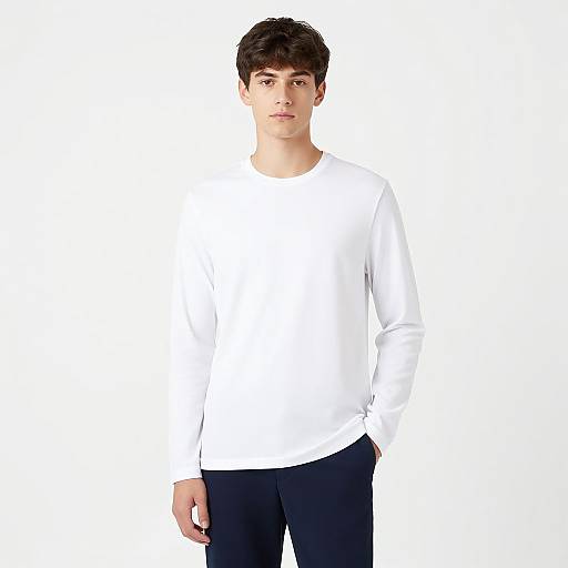 Photograph of a young man with short dark hair, wearing a plain white long-sleeve shirt and black pants, standing against a stark white background