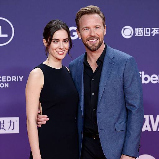 Photograph of a smiling couple at a red carpet event; woman in sleeveless black dress, man in blue blazer, black shirt, against a