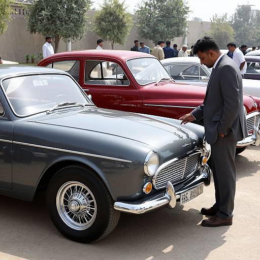 Photograph of a man in a gray suit inspecting a vintage black car with chrome details, surrounded by other classic cars, including a red one,