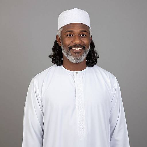 Photograph of a smiling Black man with long black hair, gray beard, wearing a white cap and long-sleeved white traditional garment, against a
