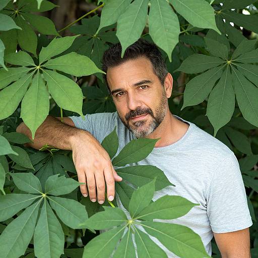 Photograph of a bearded man with dark hair, wearing a white T-shirt, partially hidden by large green leaves. He gazes directly at the