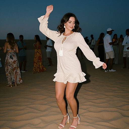 Photograph of a smiling woman in a white, long-sleeve, lace dress dancing on a sandy beach at dusk, surrounded by silhouet