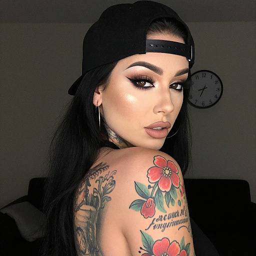 Photograph of a tattooed woman with long black hair, wearing a black backward cap, hoop earrings, and dramatic makeup, showcasing a colorful flower tattoo