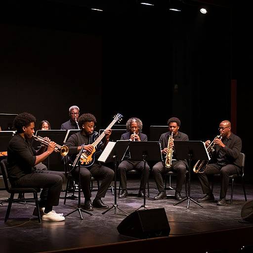 Photograph of six male jazz musicians on stage, wearing black clothes, playing saxophones and a bass guitar, seated with music stands, under dim