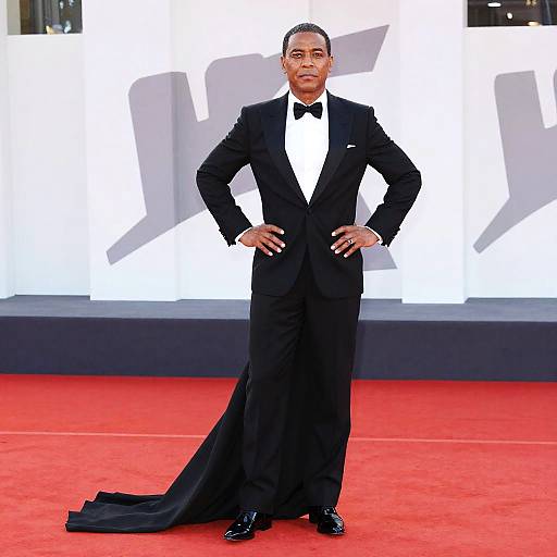 Photograph of Barack Obama in a black tuxedo with a white shirt and bow tie, standing on a red carpet, hands on hips, white