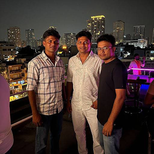 Photograph of three Indian men standing on a rooftop at night, city skyline with illuminated buildings in the background. They wear casual shirts and jeans, smiling